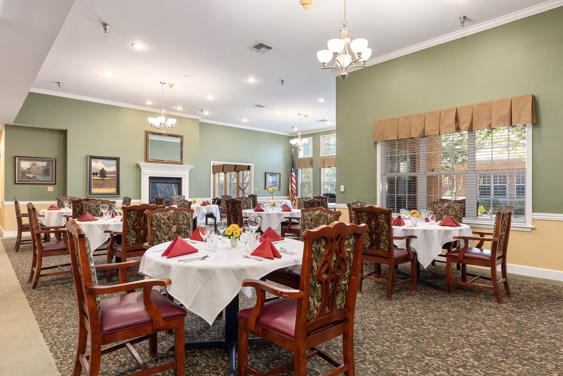 Dining room with round tables set with white tablecloths, folded red napkins, floral centerpieces and wooden chairs in a well-lit room with green walls and a fireplace.