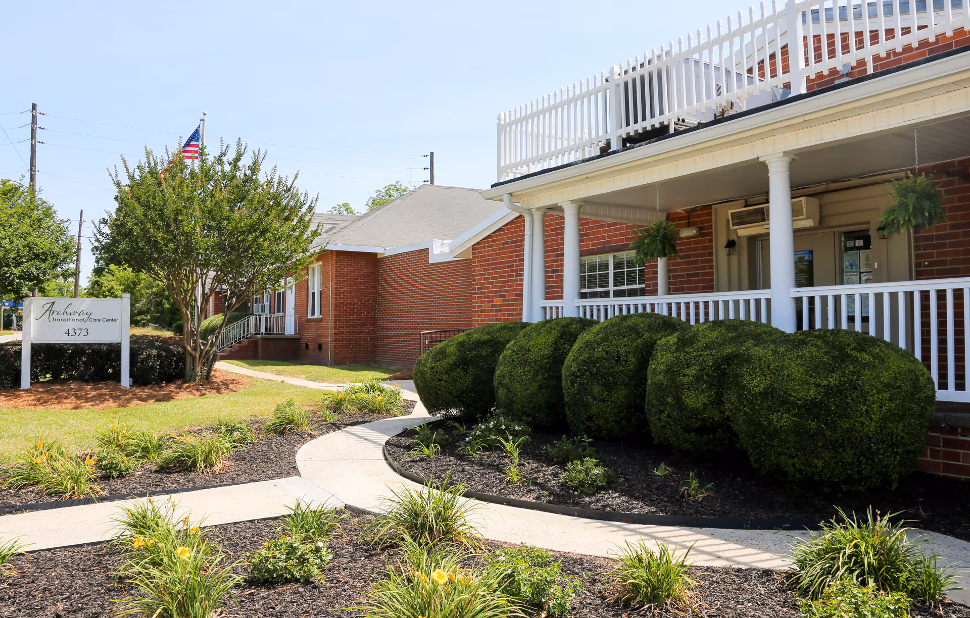 Exterior view of Archway Transitional Care facility showing a brick building with a white porch railing, neatly trimmed bushes, a curved concrete walkway, and a sign with the facility name and address 4373. There is an American flag and trees in the background under a clear blue sky.