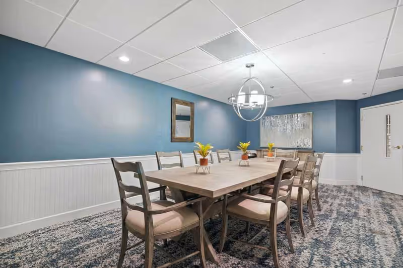 A dining room with a long rectangular wooden table surrounded by eight cushioned wooden chairs. The walls are painted blue on the upper half and white on the lower half with a chair rail. There is a modern chandelier hanging above the table, three small potted plants on the table, a mirror on one wall, and a large abstract painting on another wall. The floor is carpeted with a patterned design.