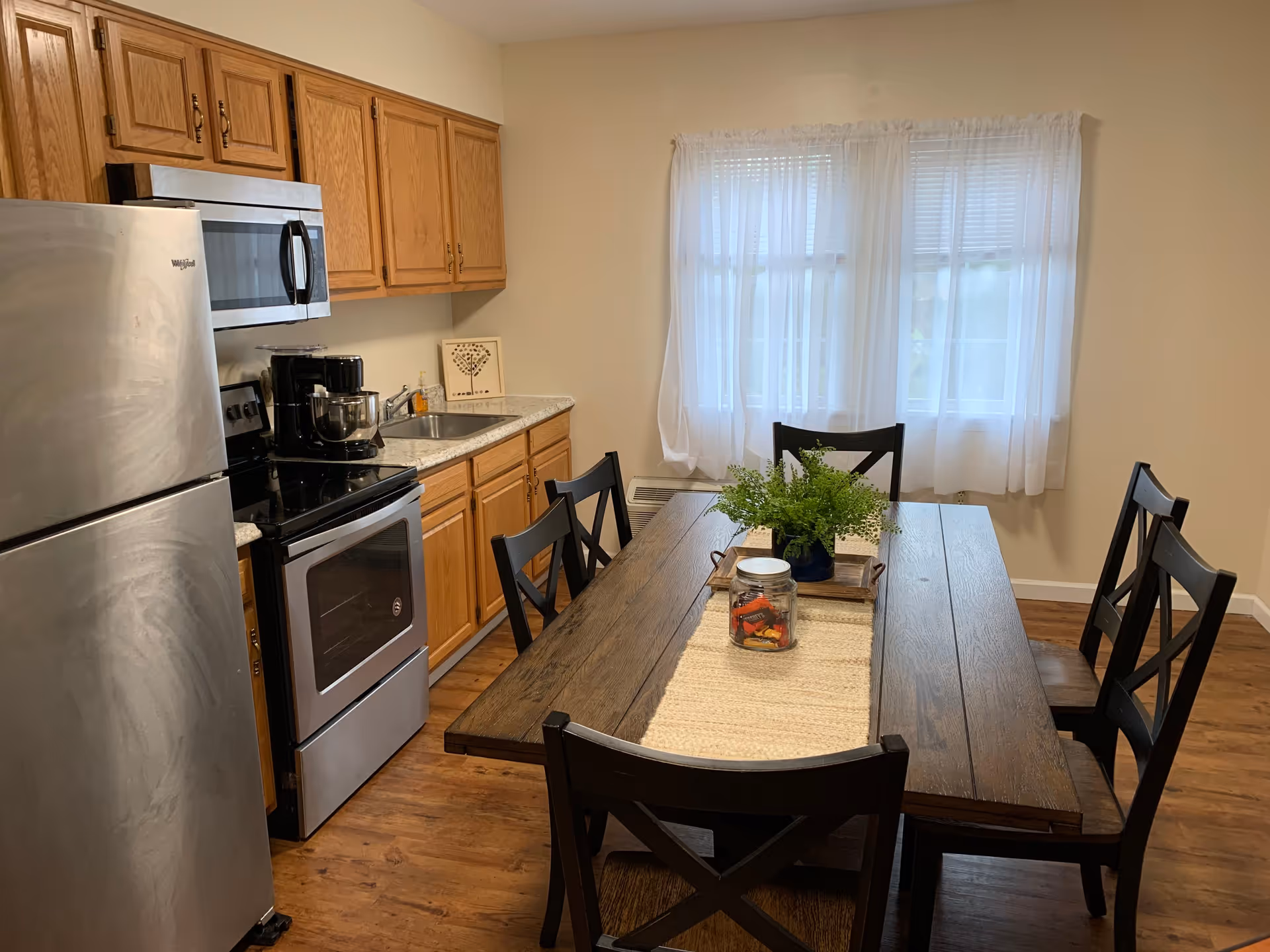 A kitchen and dining area with wooden cabinets, a stainless steel refrigerator, stove, and microwave. There is a wooden dining table with six black chairs, a beige table runner, a jar with candy, and a green plant centerpiece. A window with sheer white curtains is in the background.