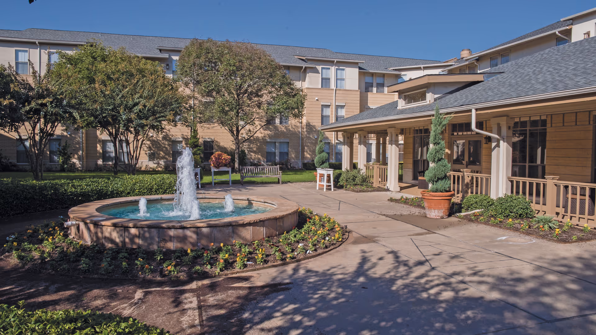 Outdoor courtyard area of Town Village Crossing featuring a circular water fountain surrounded by flowers, trees, and benches, with a multi-story building and covered porch in the background under a clear blue sky.