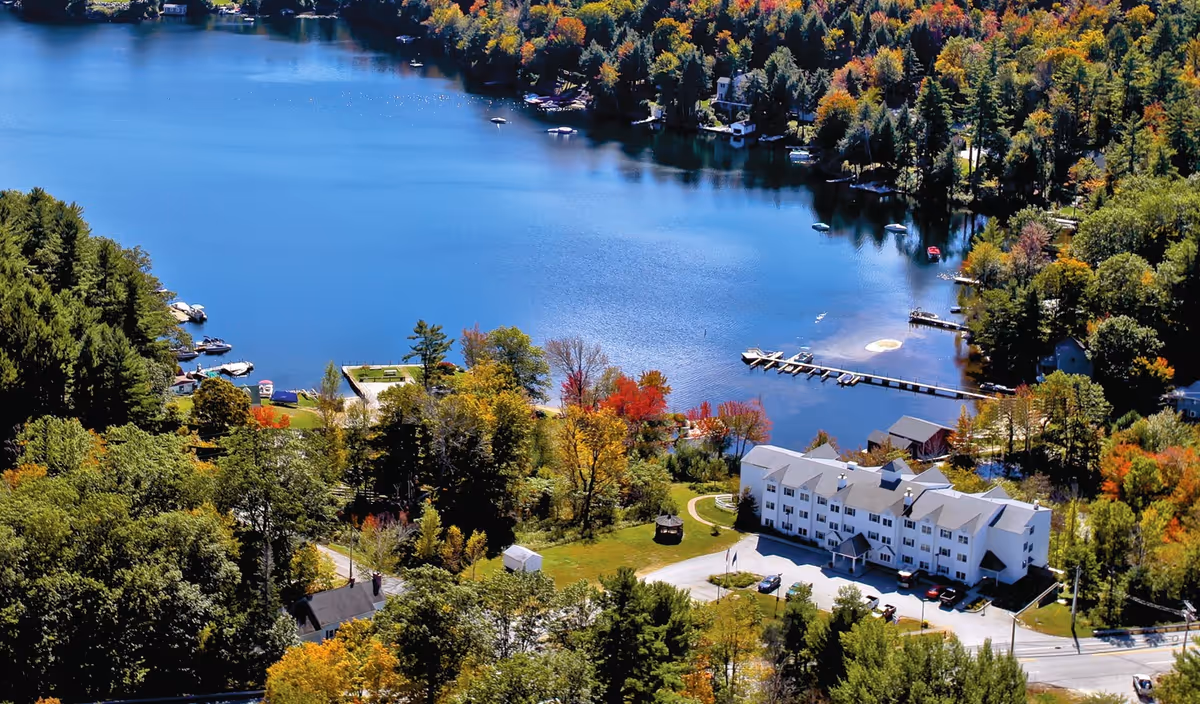 Aerial view of a large white multi-story building surrounded by trees with autumn foliage, located next to a calm blue lake with docks and boats along the shoreline.