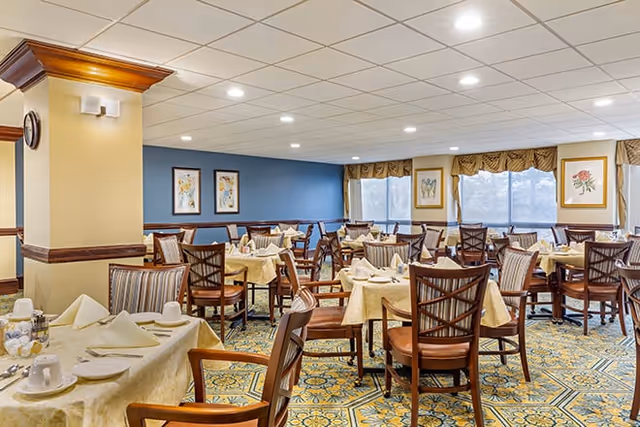 A spacious dining room with multiple tables covered in beige tablecloths, each set with folded napkins, cups, and plates. Wooden chairs with cushioned seats surround the tables. The room has a patterned carpet, blue and cream walls with framed floral artwork, and large windows with valances allowing natural light to enter.