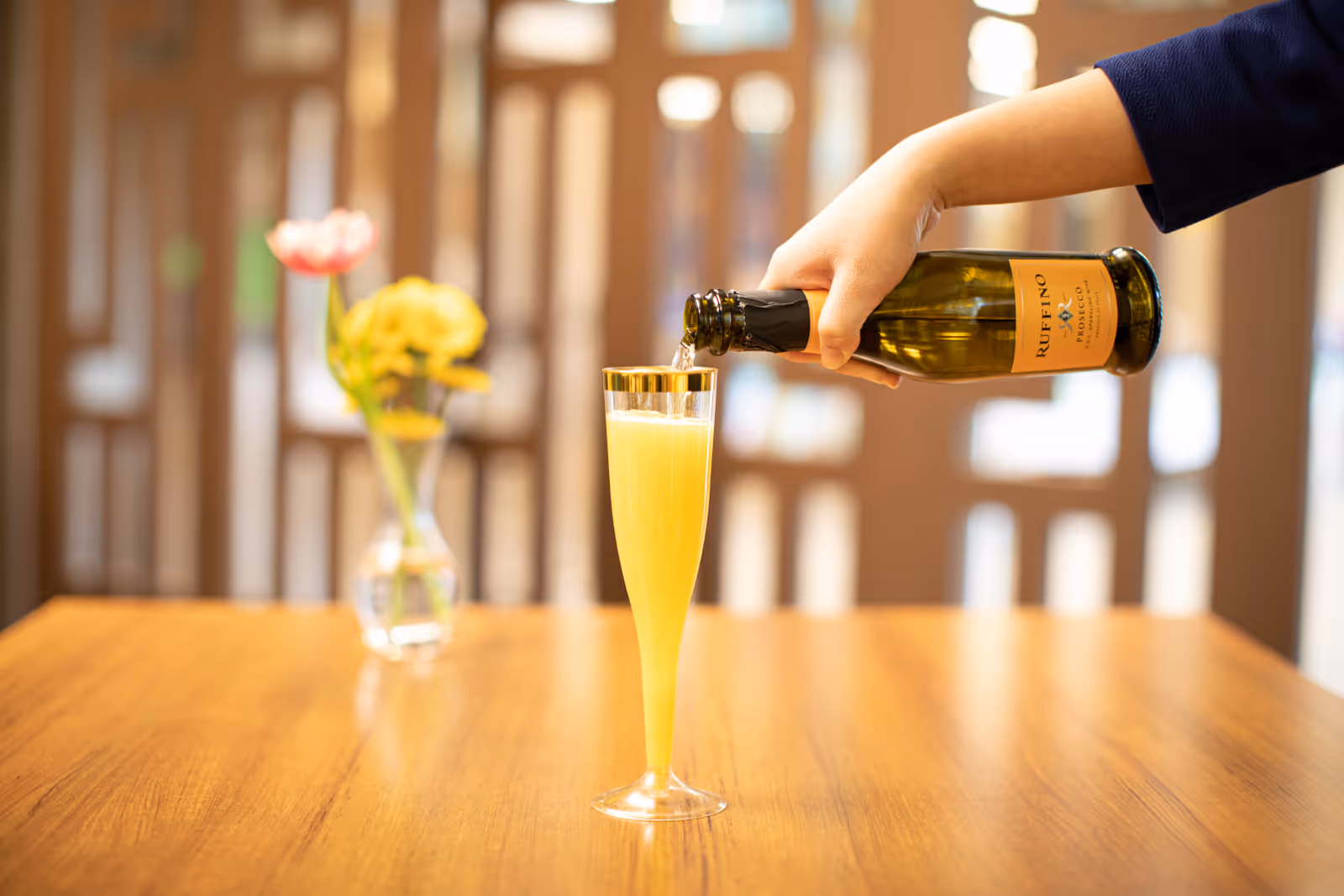 A hand is pouring a yellow beverage from a Ruffino bottle into a tall champagne flute on a wooden table. In the background, there is a blurred vase with yellow and pink flowers.