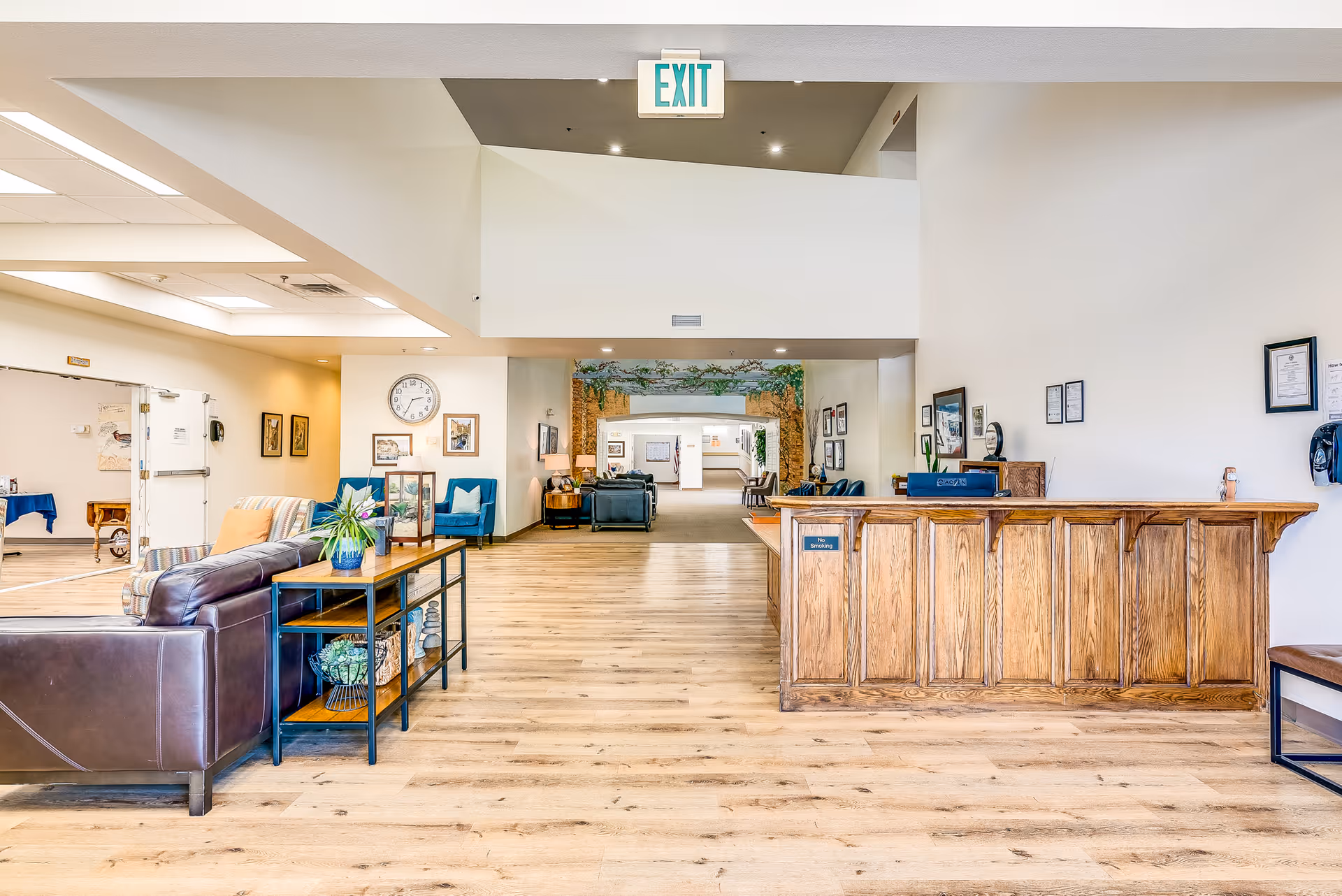 Spacious senior living facility reception lobby with a wooden front desk, seating areas, and an exit sign overhead.