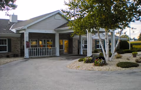 Front exterior view of Meadow View Assisted Living facility showing a covered entrance with white columns, a circular driveway, landscaped garden beds with trees and shrubs, and a partly cloudy sky.
