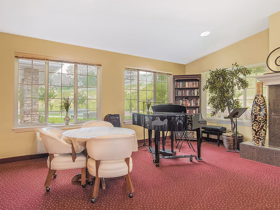 A cozy interior room with a round table covered with a white tablecloth surrounded by four cushioned chairs on a red carpet. There is a black grand piano with a music stand, a tall bookshelf filled with books, a large potted plant, and a decorative vase near a fireplace. Large windows let in natural light and show greenery outside.