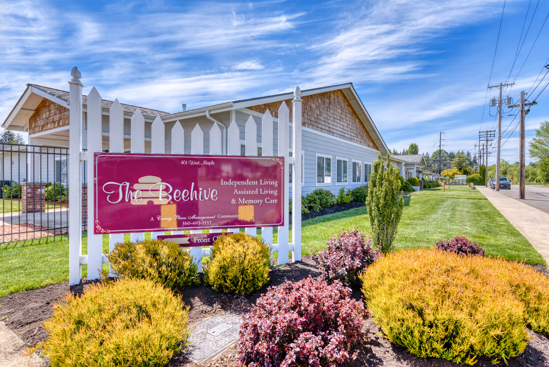 Exterior view of The Beehive Retirement Community building with a white picket fence and a maroon sign displaying the community's name and services. The building is surrounded by well-maintained landscaping with bushes and a green lawn under a partly cloudy blue sky.
