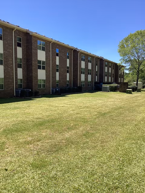 Exterior view of a three-story brick building with multiple windows, adjacent to a large grassy lawn under a clear blue sky. There is a tree on the right side and some utility boxes near the building.