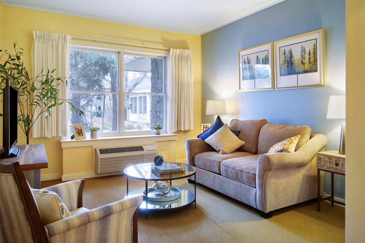 A cozy living room with a beige sofa adorned with multiple cushions, a glass coffee table with books and decorative items, a comfortable armchair, a large window with white curtains, two framed pictures of trees on a blue accent wall, and two table lamps providing warm lighting.