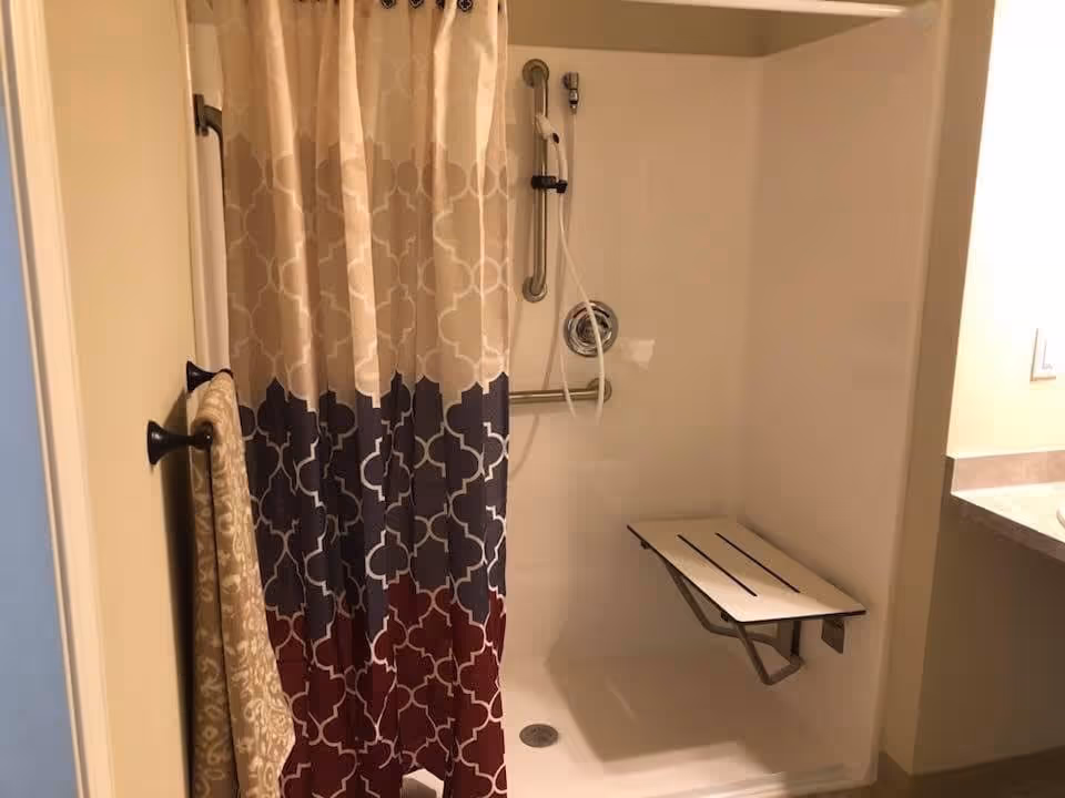 A shower area with a patterned shower curtain in beige, navy, and burgundy colors. Inside the shower, there is a foldable white shower seat attached to the wall and a handheld showerhead with a grab bar. A beige towel hangs on a black towel rack outside the shower.