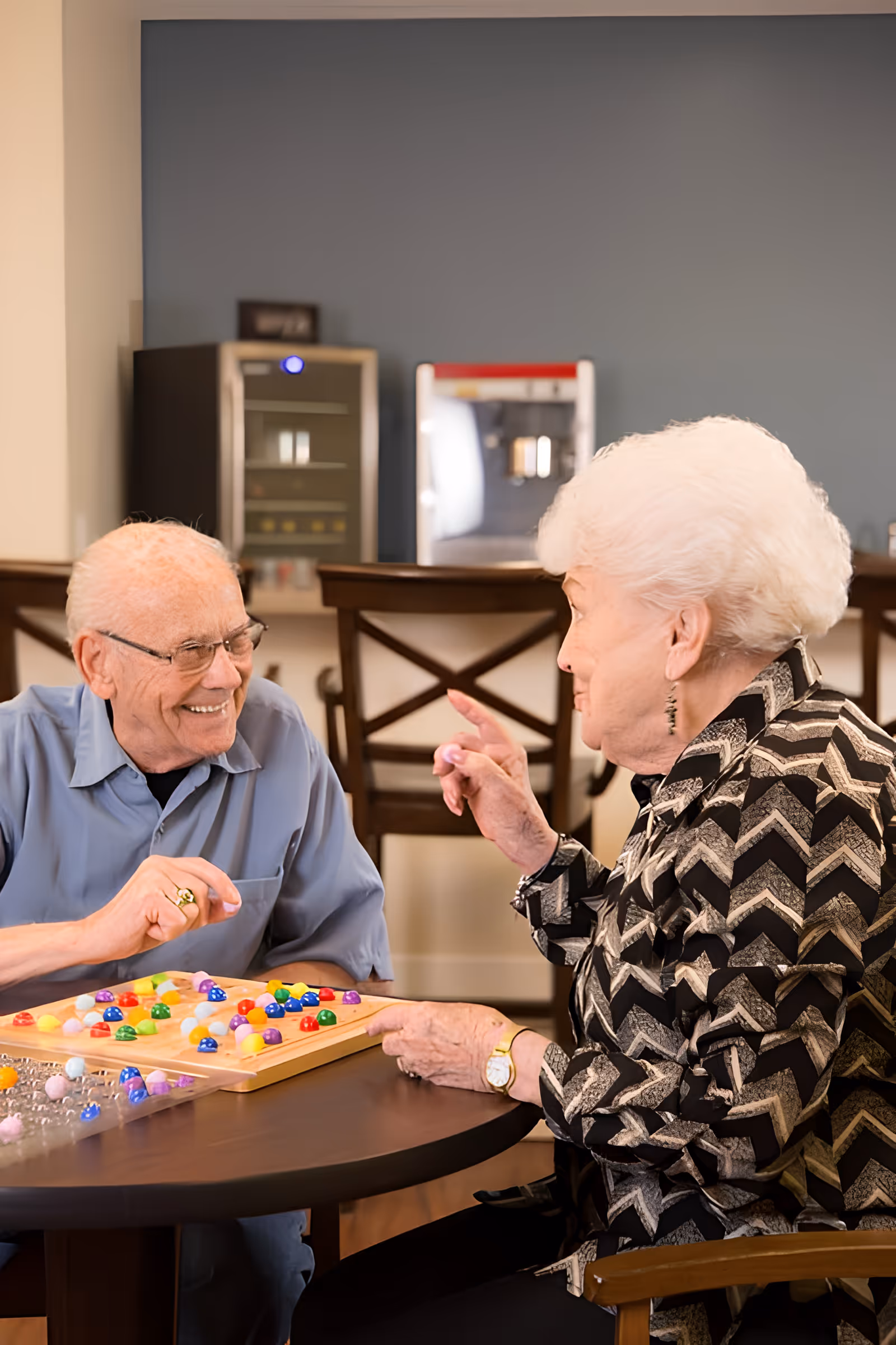 Two elderly individuals sitting at a round table playing a colorful board game, smiling and engaging with each other in a cozy indoor setting with chairs and a beverage machine in the background.