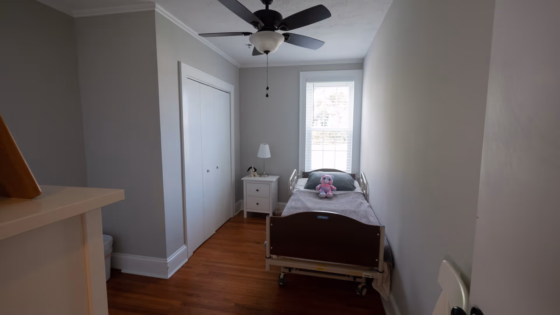 Small adjustable hospital bed in a bright bedroom with nightstand, lamp, and window.