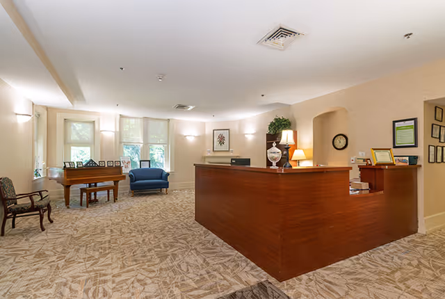 A spacious reception area with a large wooden front desk on the right side. The room has beige walls and patterned carpet flooring. On the left side, there is a wooden piano, a blue armchair, and a wooden chair. The back wall features three large windows with sheer curtains, framed pictures, and wall-mounted lights. There is a clock and framed certificates on the wall behind the desk.