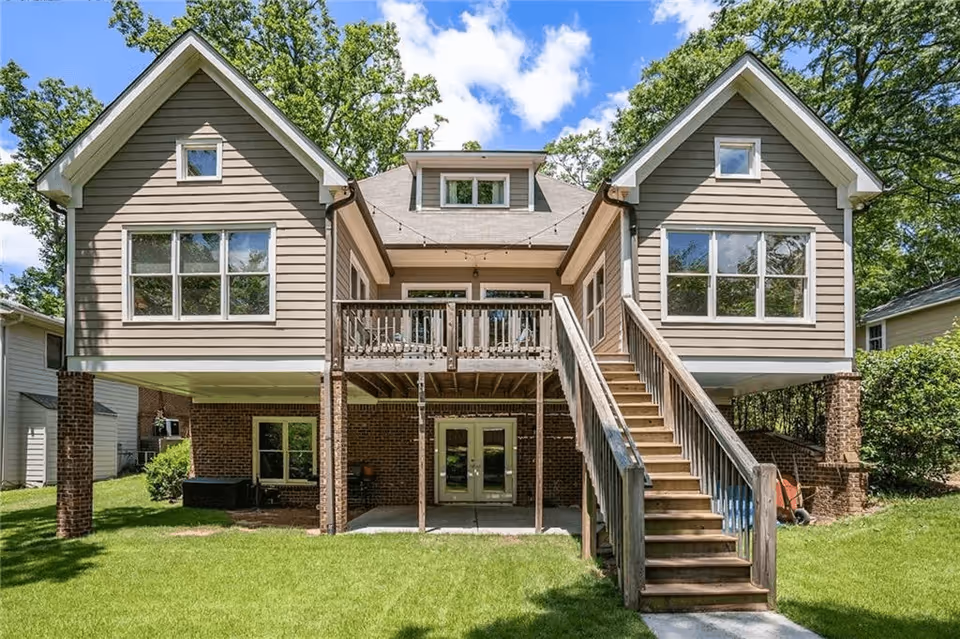 Rear exterior of a raised two-story house with twin gables, a central wooden deck and staircase leading down to a grassy yard.