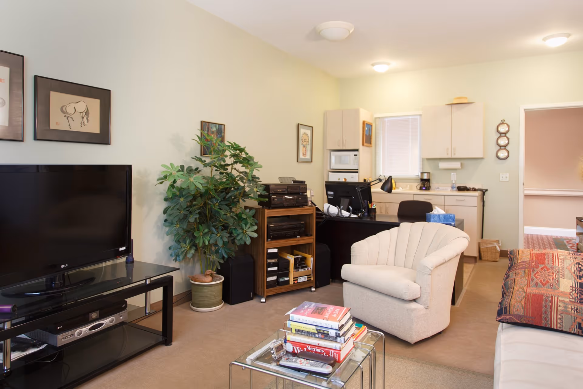 A cozy living room area in an assisted living facility featuring a beige armchair, a glass coffee table stacked with books and remote controls, a flat-screen TV on a black stand, a potted plant, and a desk with a computer and office supplies. The background shows a small kitchenette with cabinets, a microwave, and a coffee maker.