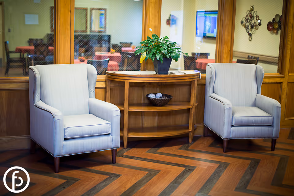 Two upholstered armchairs flank a wooden console table with a potted plant in a seating area with wood-patterned flooring and windowed wall panels.
