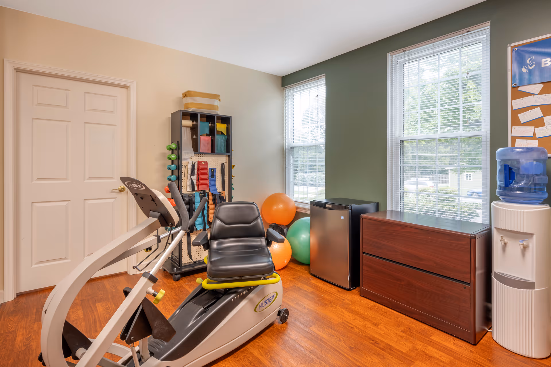 A small exercise room with a recumbent exercise bike, a rack holding various weights and resistance bands, two large exercise balls, a mini refrigerator, a wooden cabinet, and a water cooler. The room has wooden flooring, two large windows with blinds, and walls painted in beige and green.