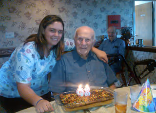 A smiling elderly man sitting at a table with a birthday cake in front of him, decorated with lit candles showing the number 98. A woman in a floral scrub top is leaning in next to him with her arm around his shoulder. Another elderly man is seated in the background near a walker. The setting appears to be a decorated room with floral wallpaper.