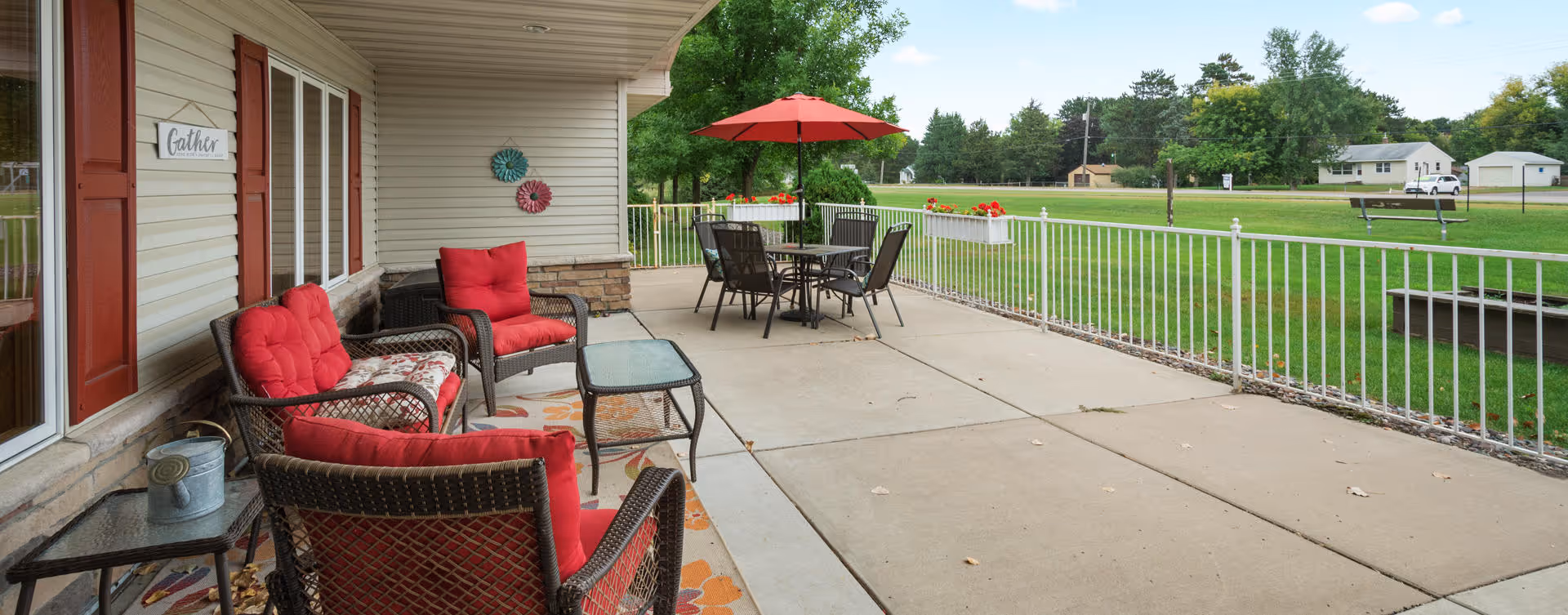 Outdoor patio area at Encore Assisted Living and Memory Care at Hugo with red cushioned wicker chairs, a glass-top table, a dining table with chairs under a red umbrella, and a white railing overlooking a green lawn and trees.