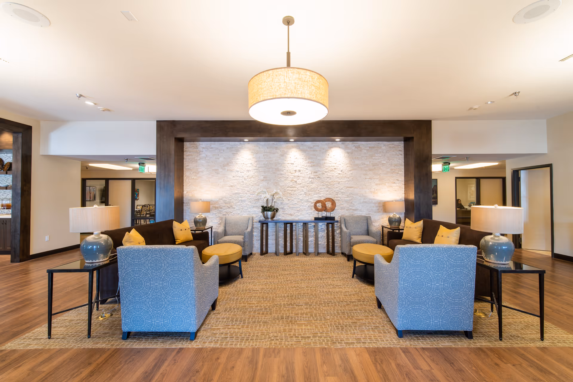 A spacious and well-lit living room area in an assisted living facility featuring two brown sofas with yellow pillows, four blue patterned armchairs, two round yellow ottomans, and several side tables with blue ceramic lamps. The back wall has a textured white stone finish with decorative plants and sculptures on black tables. The floor is wood with a large area rug, and a large circular ceiling light fixture hangs above.
