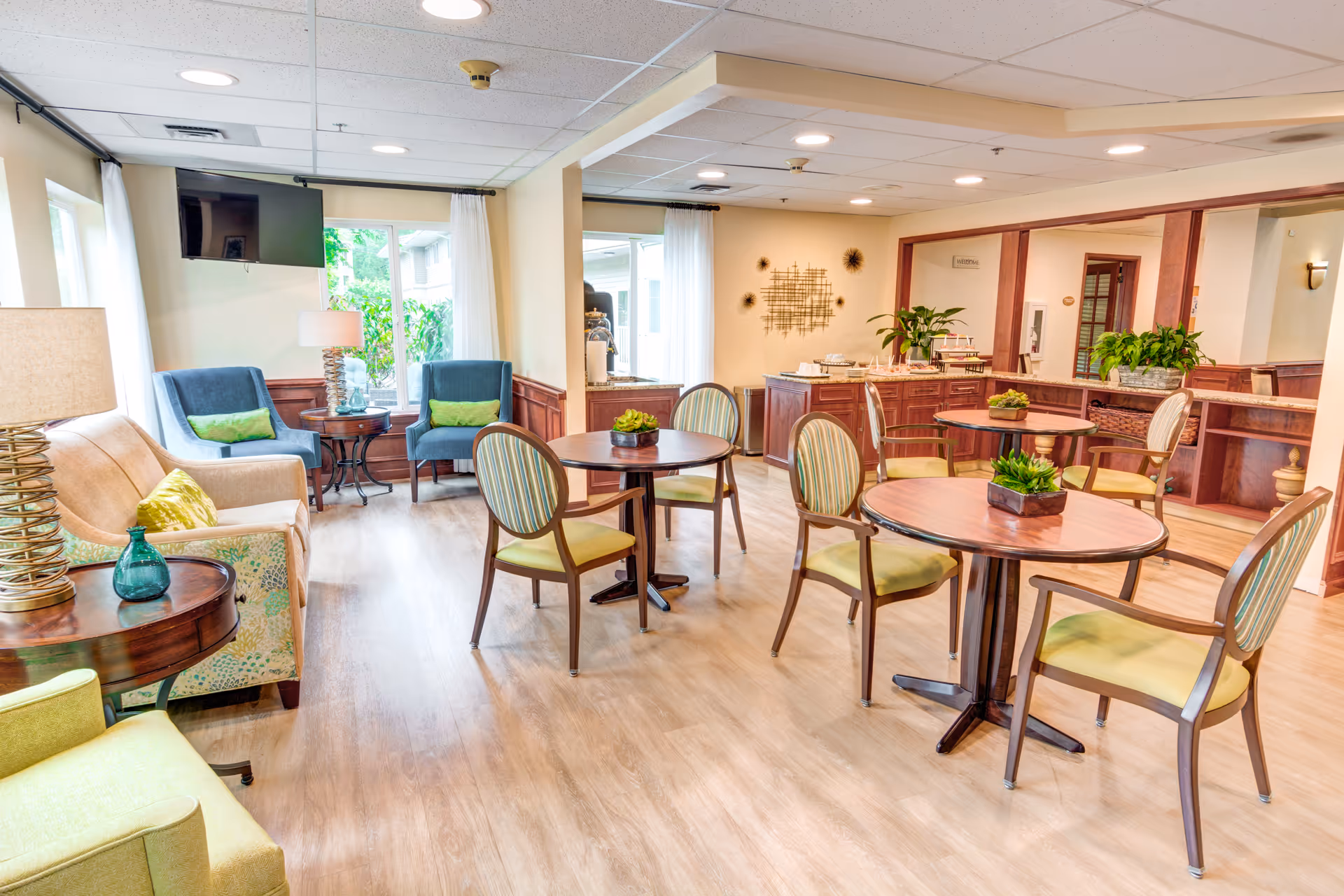 A bright and spacious senior living common area with several round wooden tables surrounded by striped cushioned chairs. There are two blue armchairs and a beige sofa with green pillows near large windows letting in natural light. The room features wood paneling, a mounted TV, decorative plants on tables, and a buffet counter with additional plants and decor.