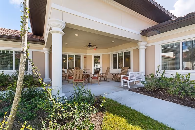 Covered outdoor patio area at Discovery Village At Palm Beach Gardens with several wooden chairs and a table, surrounded by beige walls with white columns and windows, and landscaped greenery in the foreground.