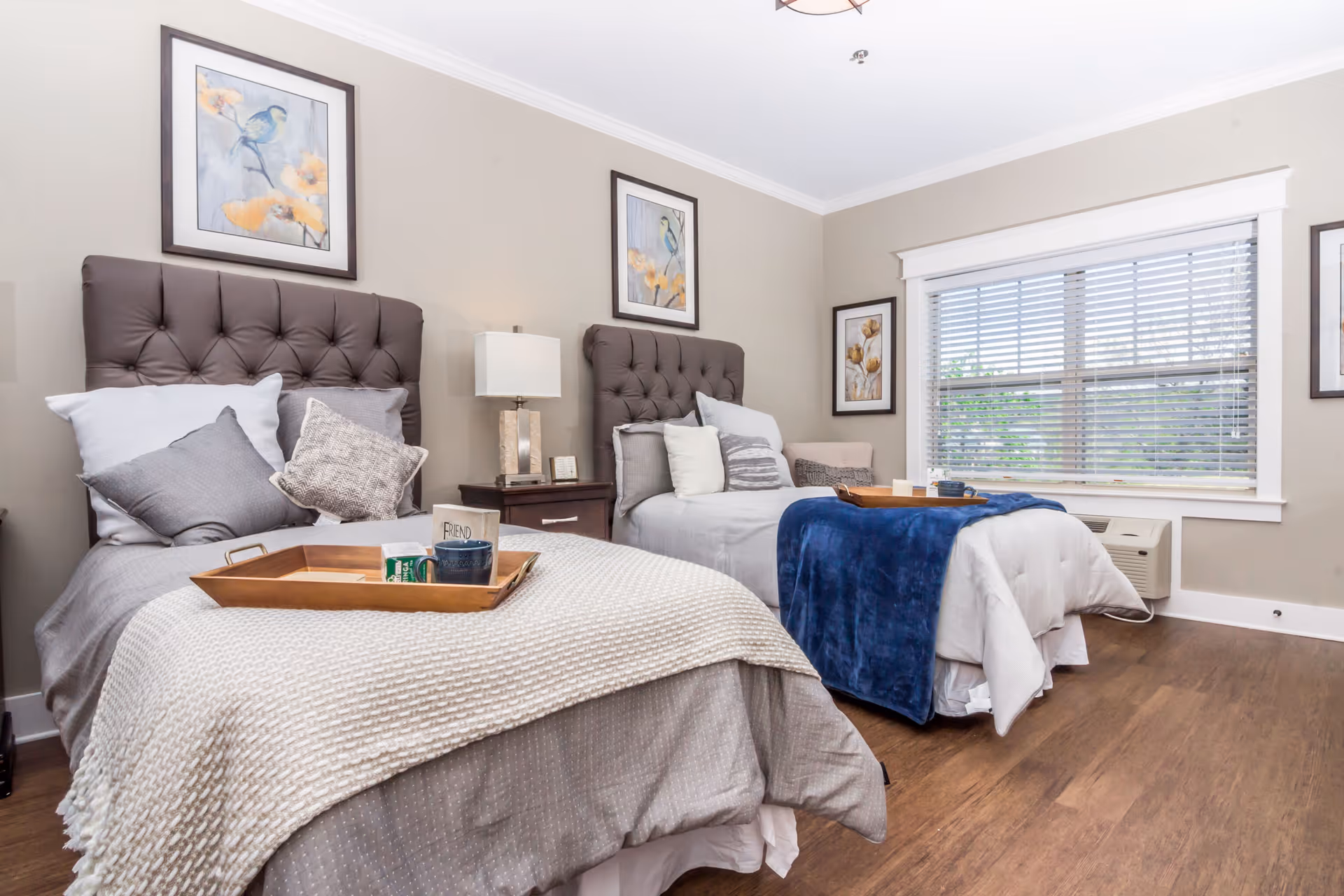 A bright and cozy bedroom with two twin beds featuring tufted headboards. Each bed is neatly made with pillows and blankets, one with a beige knitted throw and the other with a blue blanket. A wooden nightstand with a lamp and small decor items sits between the beds. The room has light beige walls adorned with framed bird and floral artwork. A large window with white blinds lets in natural light, and the floor is a warm wood tone.