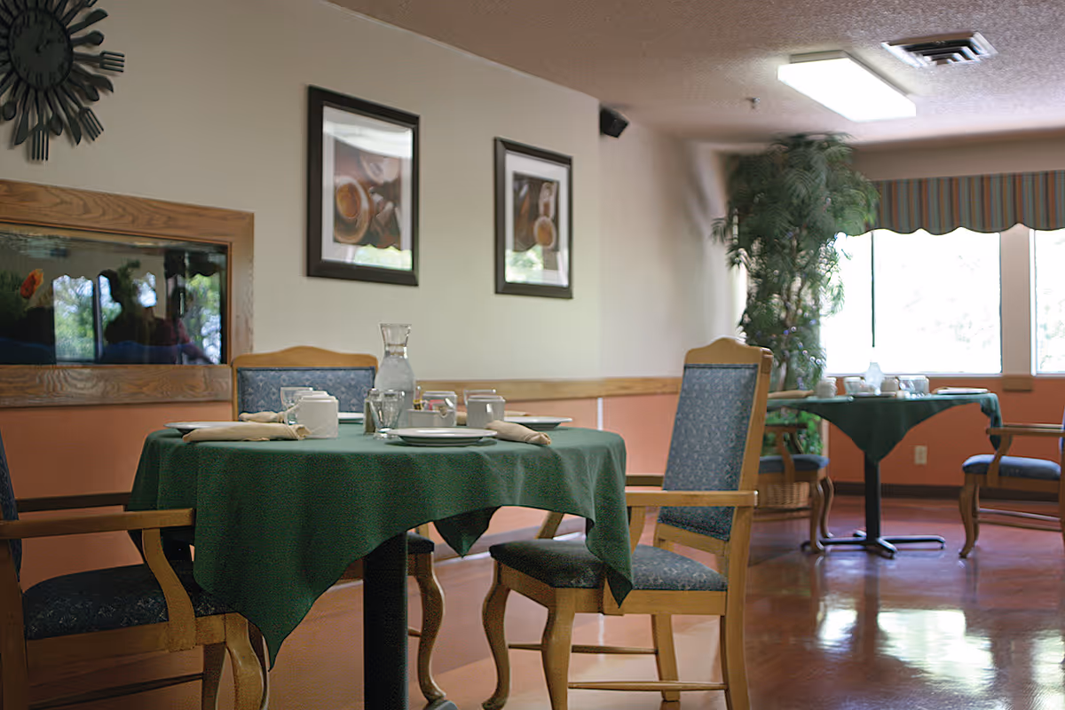 A dining room with round tables covered in green tablecloths, set with plates, cups, napkins, and a water pitcher. Wooden chairs with blue cushioned seats surround the tables. The room has framed pictures on the wall, a large window with striped valance, and a tall potted plant in the corner.
