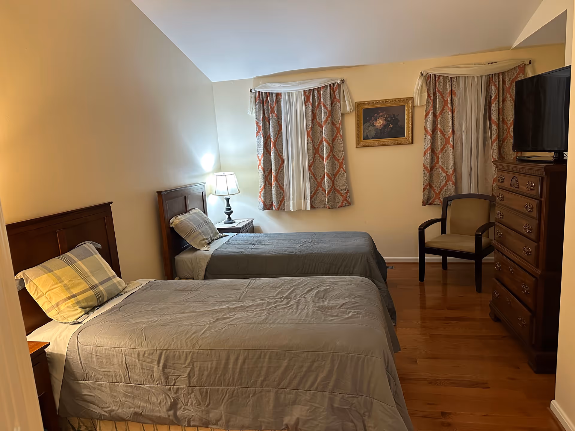 A bedroom with two single beds covered in gray bedspreads and plaid pillows. There is a wooden nightstand with a lamp between the beds. The room has two windows with patterned curtains and a framed floral painting on the wall. A wooden dresser with a television on top and a cushioned chair are also visible. The floor is wooden.