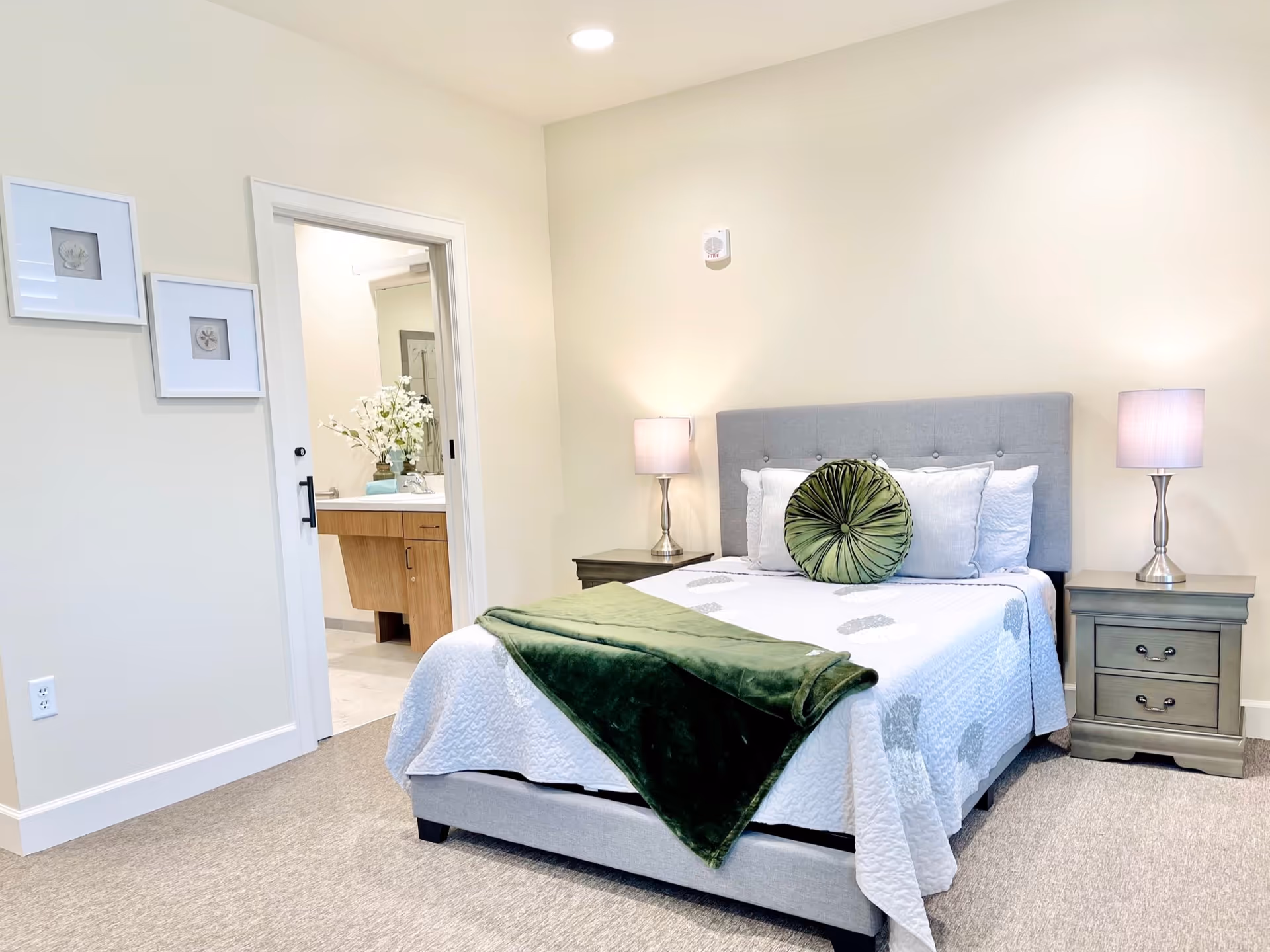 A neatly made bed with a gray upholstered headboard, white bedding with gray patterns, and a green round decorative pillow and matching green throw blanket. There are two matching nightstands with silver lamps on either side of the bed. The room has beige walls and carpeted floor. A doorway leads to a bathroom with a wooden vanity and a vase of white flowers.