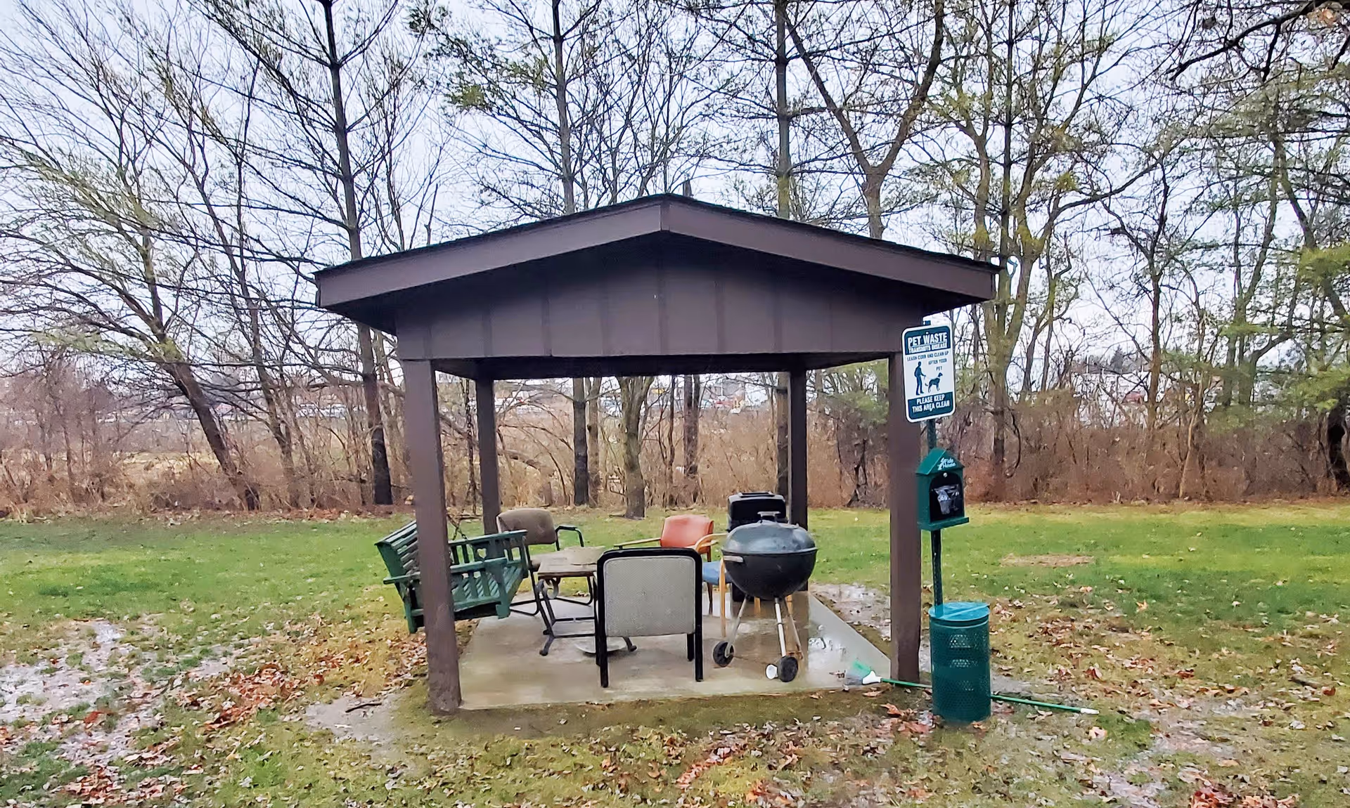 A small outdoor pavilion with a roof supported by four posts, containing several chairs and a green bench swing. There is a charcoal grill and a pet waste station with a sign nearby. The pavilion is situated on a grassy area with trees in the background.