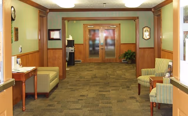 Interior hallway of a senior living facility with green walls and wood paneling. There are two armchairs and a small table on the right side, and a small desk with a chair on the left side. Double wooden doors with frosted glass are at the end of the hallway, and a water cooler is visible near the doors.