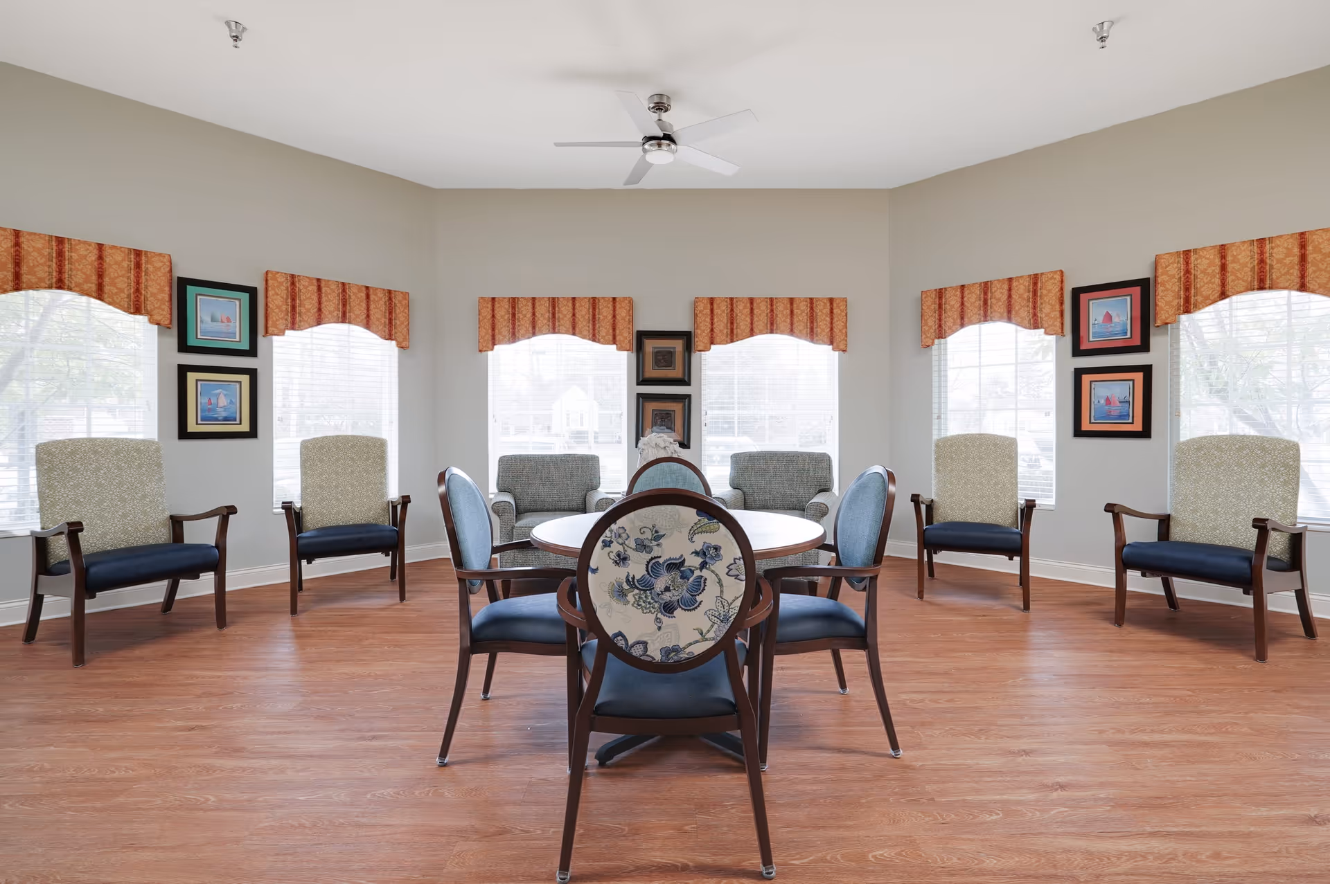 A bright and spacious sitting area with multiple chairs arranged around a circular table in the center and additional chairs lined up against the walls. The room has large windows with red and orange patterned valances, light-colored walls, and wood flooring. Several framed pictures hang on the walls between the windows.