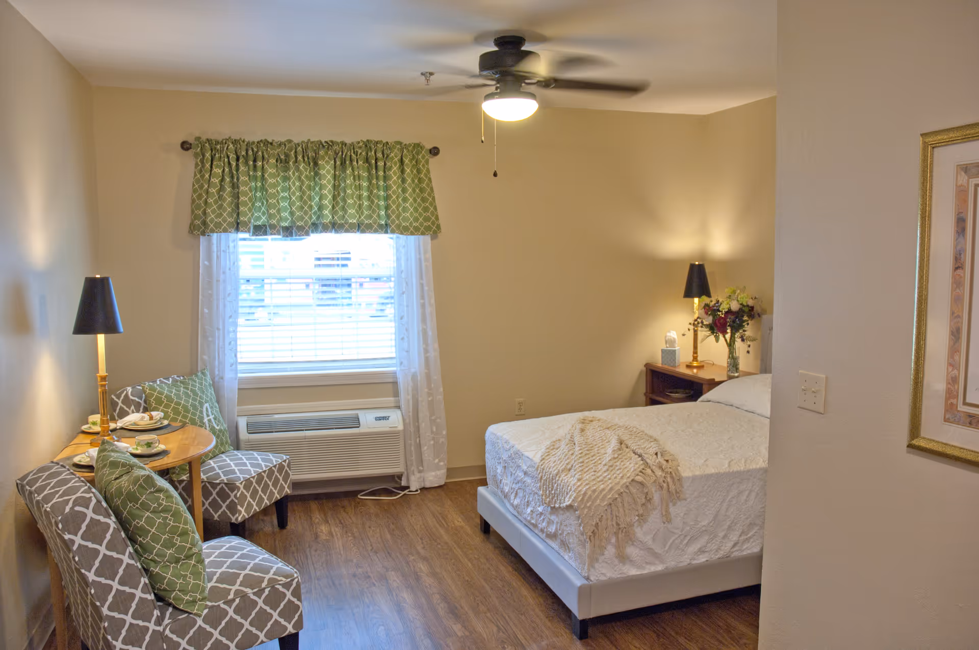 A cozy senior living bedroom with a bed covered in a white textured bedspread and a beige throw blanket. There are two patterned chairs with green and white pillows next to a small round wooden table set with teacups. A window with green and white curtains is above an air conditioning unit. The room has wooden flooring, a ceiling fan with a light, two bedside tables with lamps, and a framed picture on the wall.
