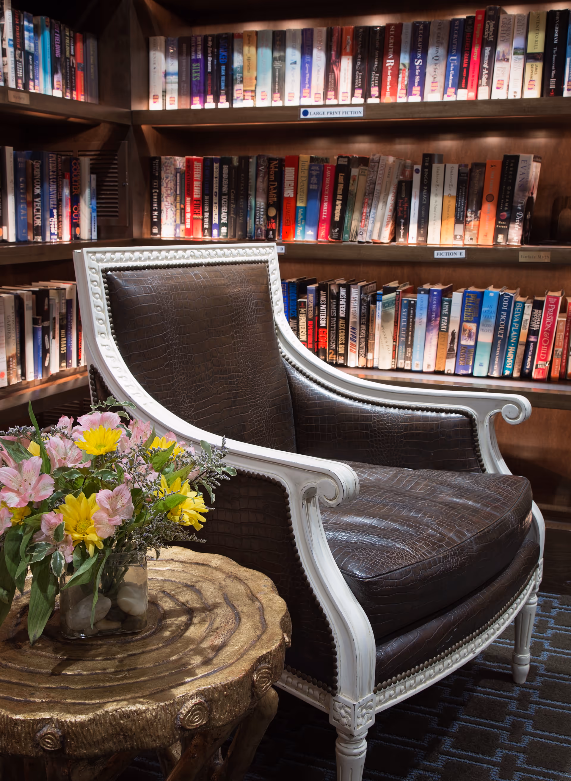 A cozy reading nook featuring a dark brown leather armchair with white wooden frame and nailhead trim, positioned next to a rustic wooden side table holding a glass vase with a colorful bouquet of yellow and pink flowers. Behind the chair, there are shelves filled with a variety of books organized by category labels such as 'Large Print Fiction' and 'Fiction E'.