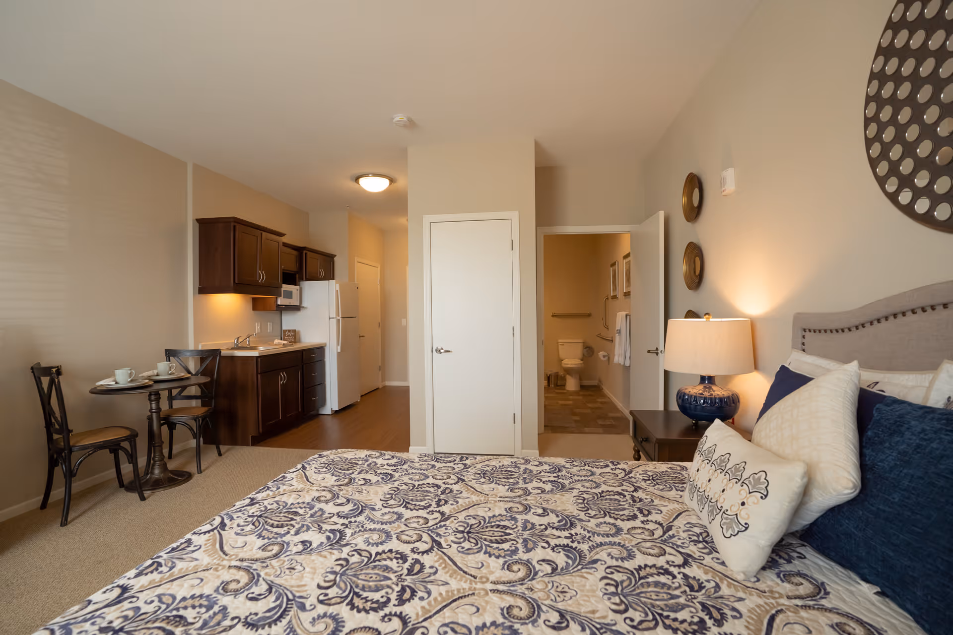 A cozy studio apartment featuring a bed with patterned bedding and multiple pillows, a bedside table with a lamp, a small dining table with two chairs and cups, a kitchenette with dark wood cabinets, a refrigerator, and a microwave. In the background, there is an open door leading to a bathroom with a visible toilet and towel rack.