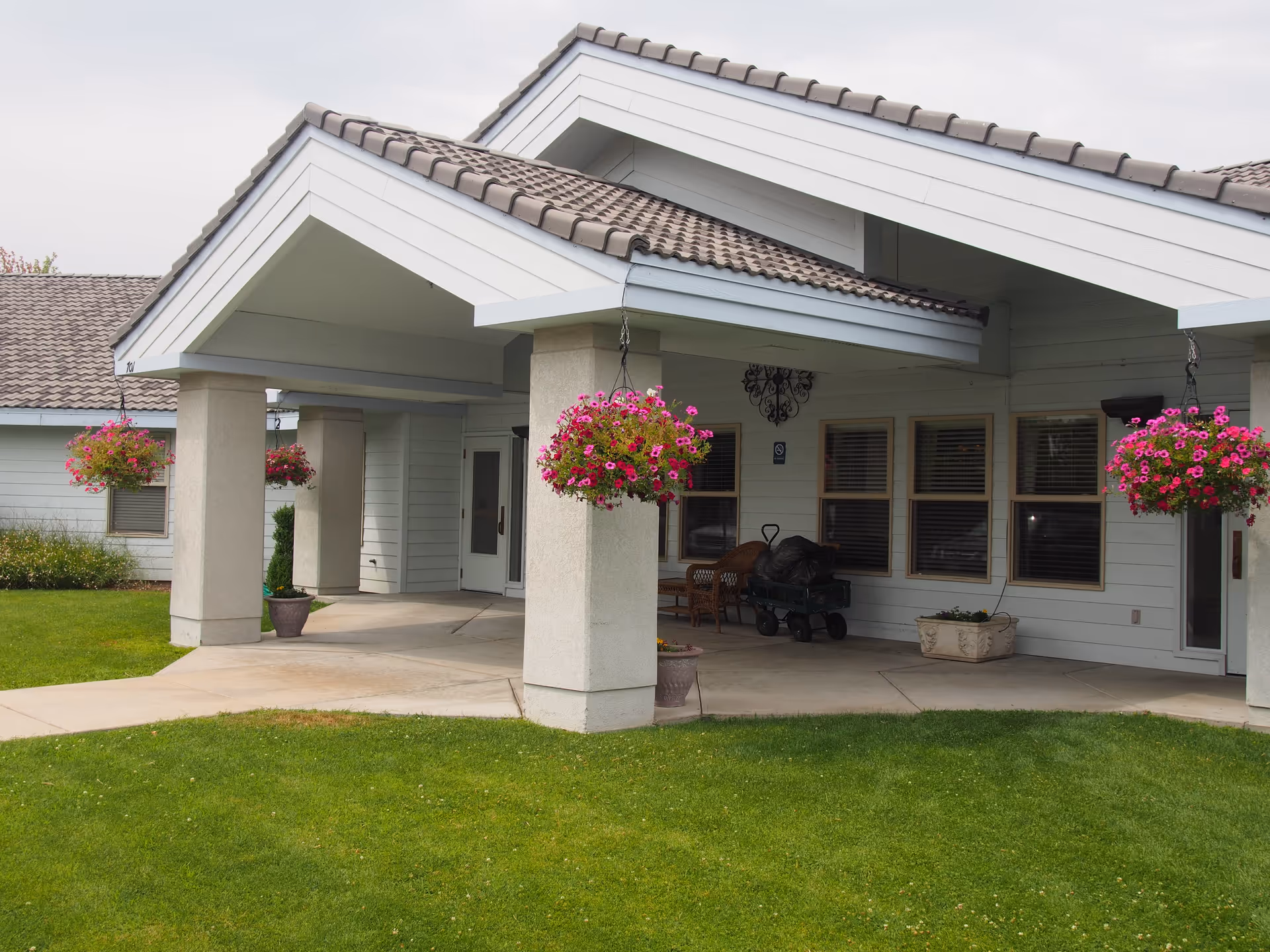 Entrance of a building with a covered porch supported by columns, hanging flower baskets with pink flowers, a green lawn in front, and windows along the wall under a tiled roof.
