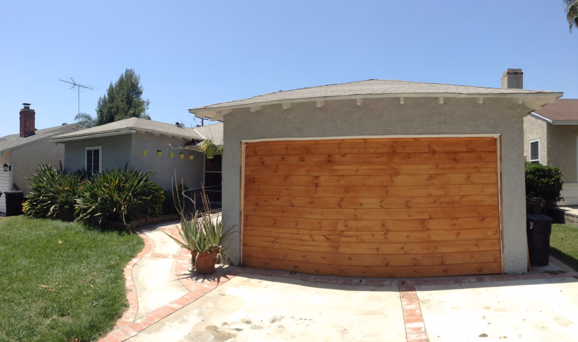 Exterior view of a single-story building with a wooden garage door, a concrete driveway with brick accents, green lawn, and various plants including a potted aloe near the driveway. The sky is clear and blue.