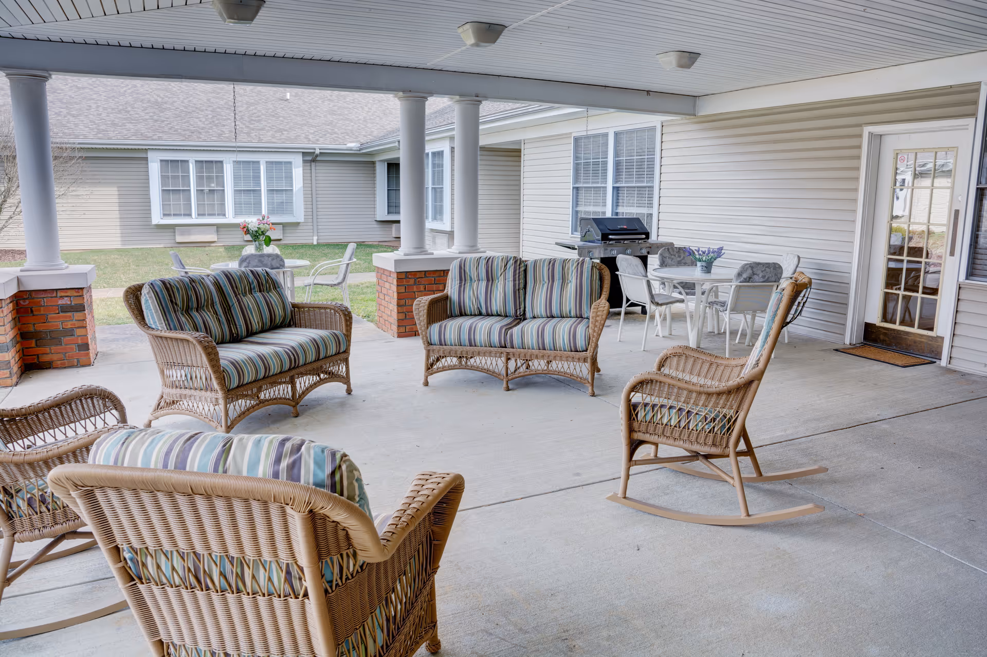 Covered outdoor patio area with wicker furniture including two loveseats, two rocking chairs with striped cushions, a round table with chairs, a barbecue grill, and a view of the building exterior with windows and a door.