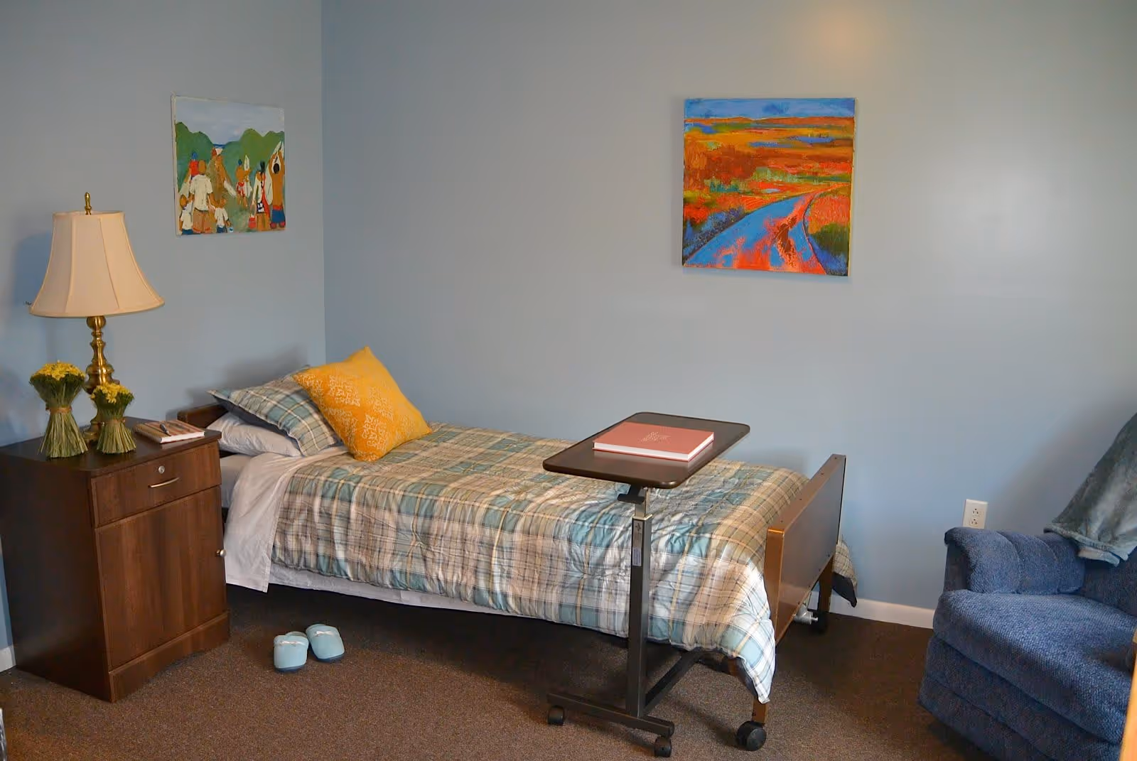 A small bedroom with a single bed covered in a plaid bedspread and a yellow pillow. Next to the bed is a wooden nightstand with a lamp and two small vases with yellow flowers. There are two colorful paintings on the light blue walls. A blue armchair with a blanket draped over the back is positioned near the bed. A small overbed table with a closed book sits on the bed. A pair of light blue slippers are on the carpeted floor beside the bed.
