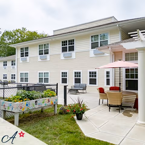 Outdoor patio area of a senior living facility with a table, chairs, and a pink umbrella. There is a raised garden bed with colorful handprints painted on its sides and some potted plants nearby. The building has beige siding with multiple windows and air conditioning units.