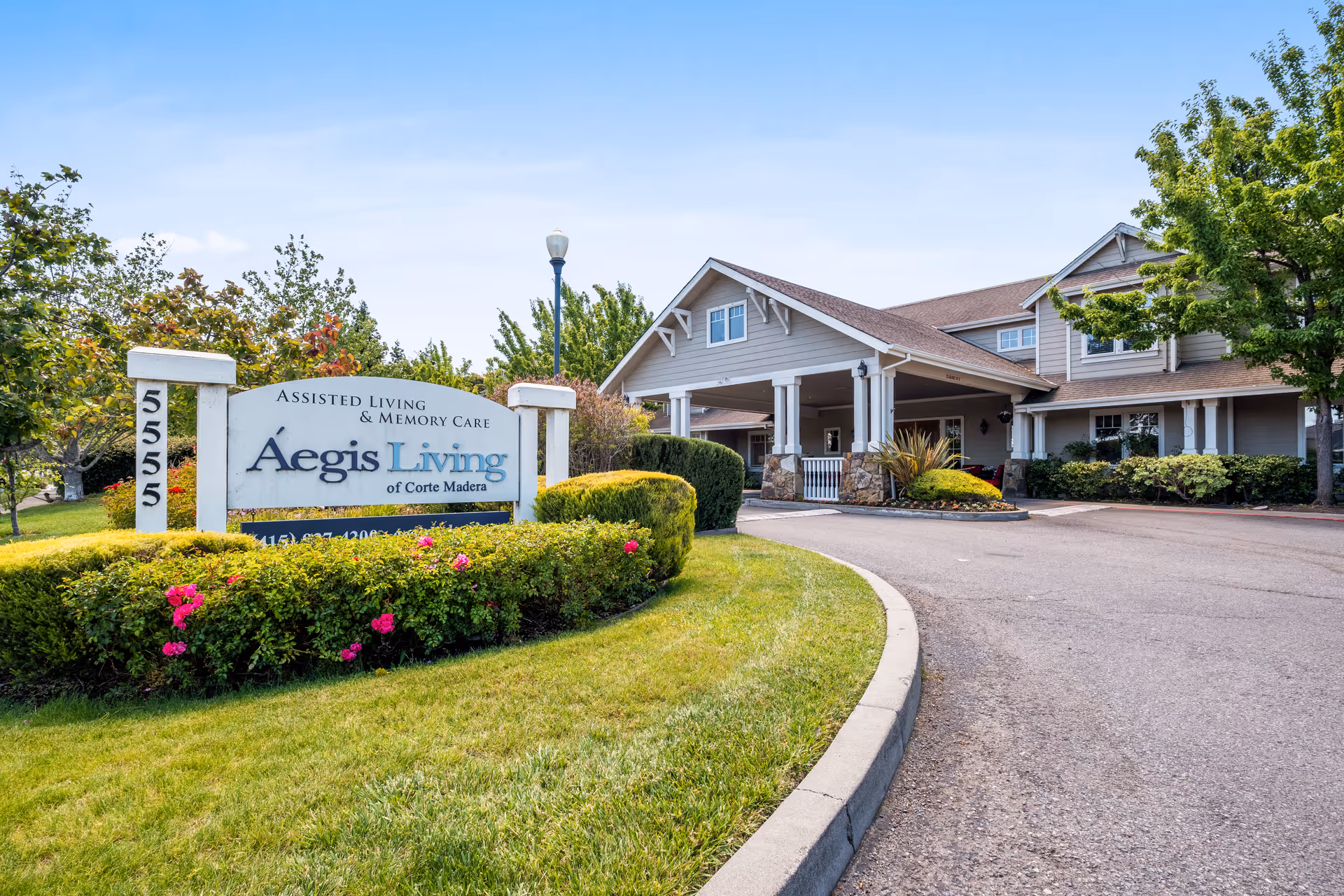 Exterior view of Aegis Living of Corte Madera assisted living and memory care facility with a driveway, landscaped greenery, and a sign displaying the facility name and address number 5555.