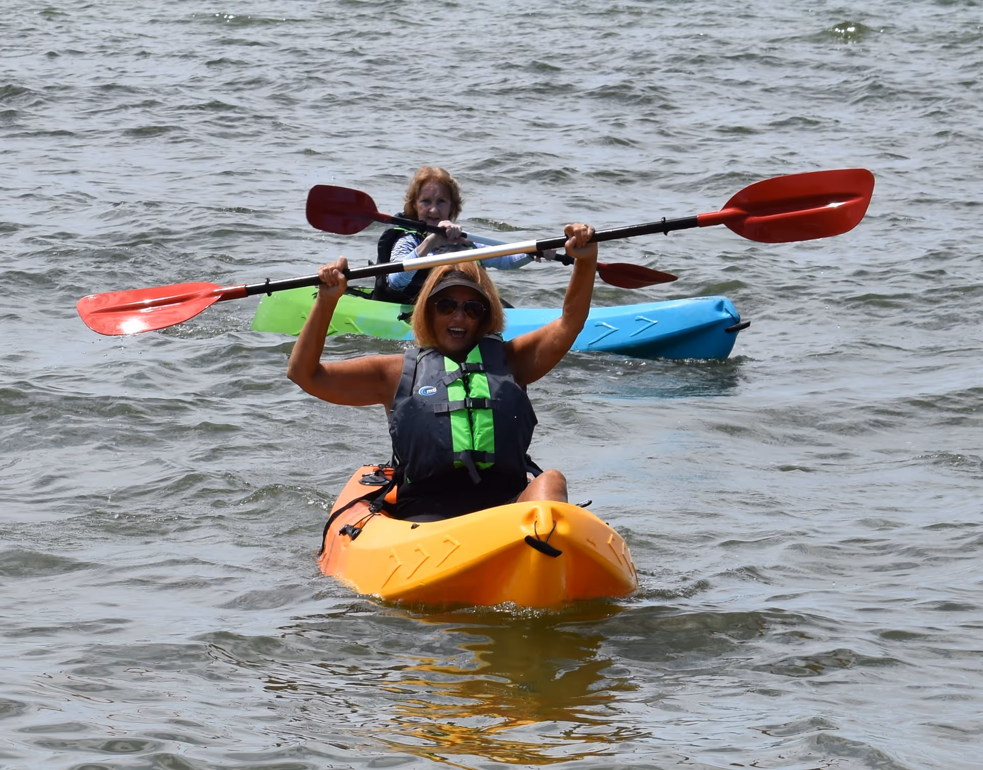 Two women kayaking on a body of water. The woman in the foreground is in an orange kayak, wearing a green and black life jacket and sunglasses, holding a paddle above her head. The woman behind her is in a blue kayak, also holding a paddle.
