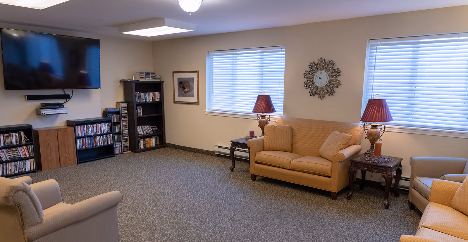 A cozy living room in a senior living community featuring beige upholstered sofas and armchairs, two side tables with decorative lamps, a wall clock, two windows with blinds, a large flat-screen TV mounted on the wall, and several bookshelves filled with DVDs and books.