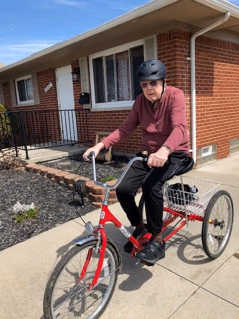 An elderly man wearing a black helmet and sunglasses rides a red adult tricycle on a sidewalk in front of a brick house with white trim and a small garden bed.