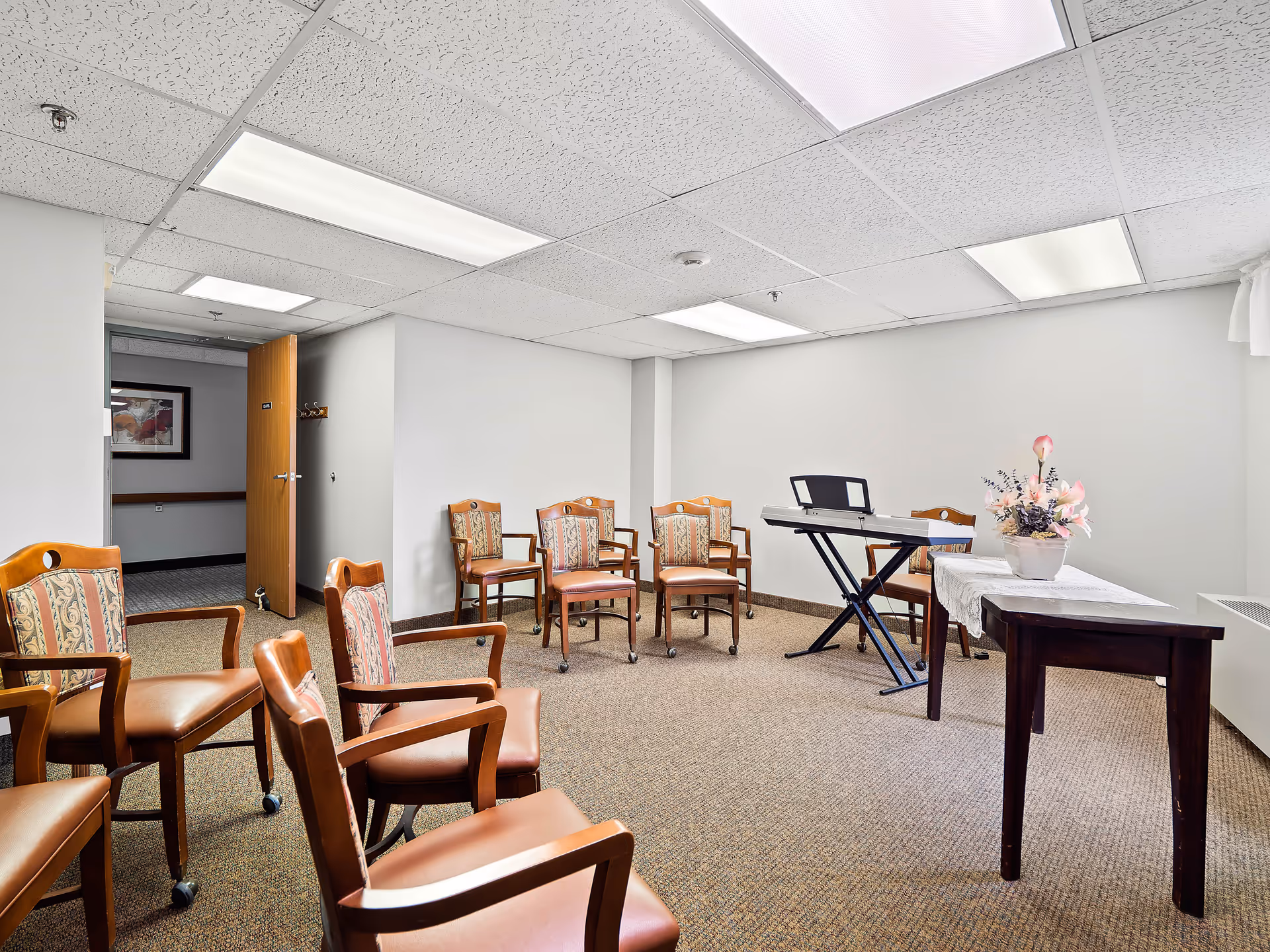 A small meeting or activity room with several wooden chairs arranged in a semi-circle. There is a keyboard on a stand and a table with a white lace cloth and a floral arrangement. The room has white walls, a carpeted floor, and a drop ceiling with fluorescent lights. An open wooden door leads to a hallway with a framed picture on the wall.