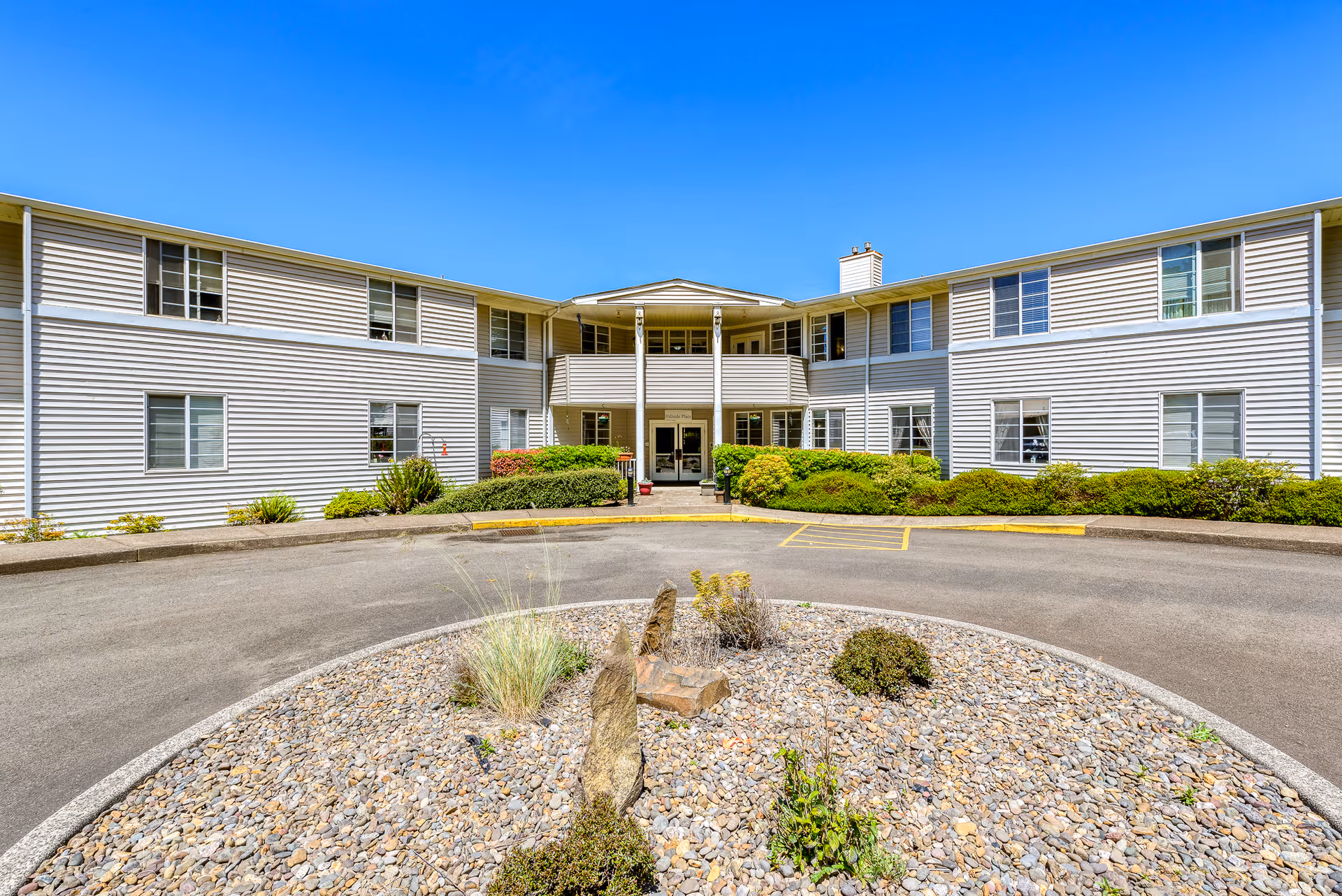 Exterior view of a two-story assisted living facility building with white siding, multiple windows, and a covered entrance. In front of the building is a circular driveway with a landscaped rock garden in the center, featuring some plants and shrubs under a clear blue sky.