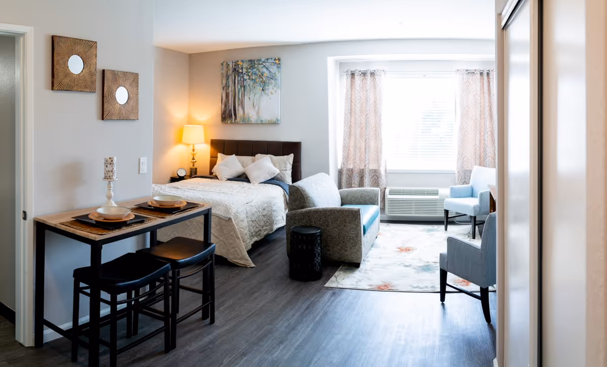 A bright and cozy senior living studio apartment featuring a bed with pillows and a bedside lamp, a small dining table set for two, a patterned sofa, two armchairs, a colorful area rug, and a large window with curtains letting in natural light.