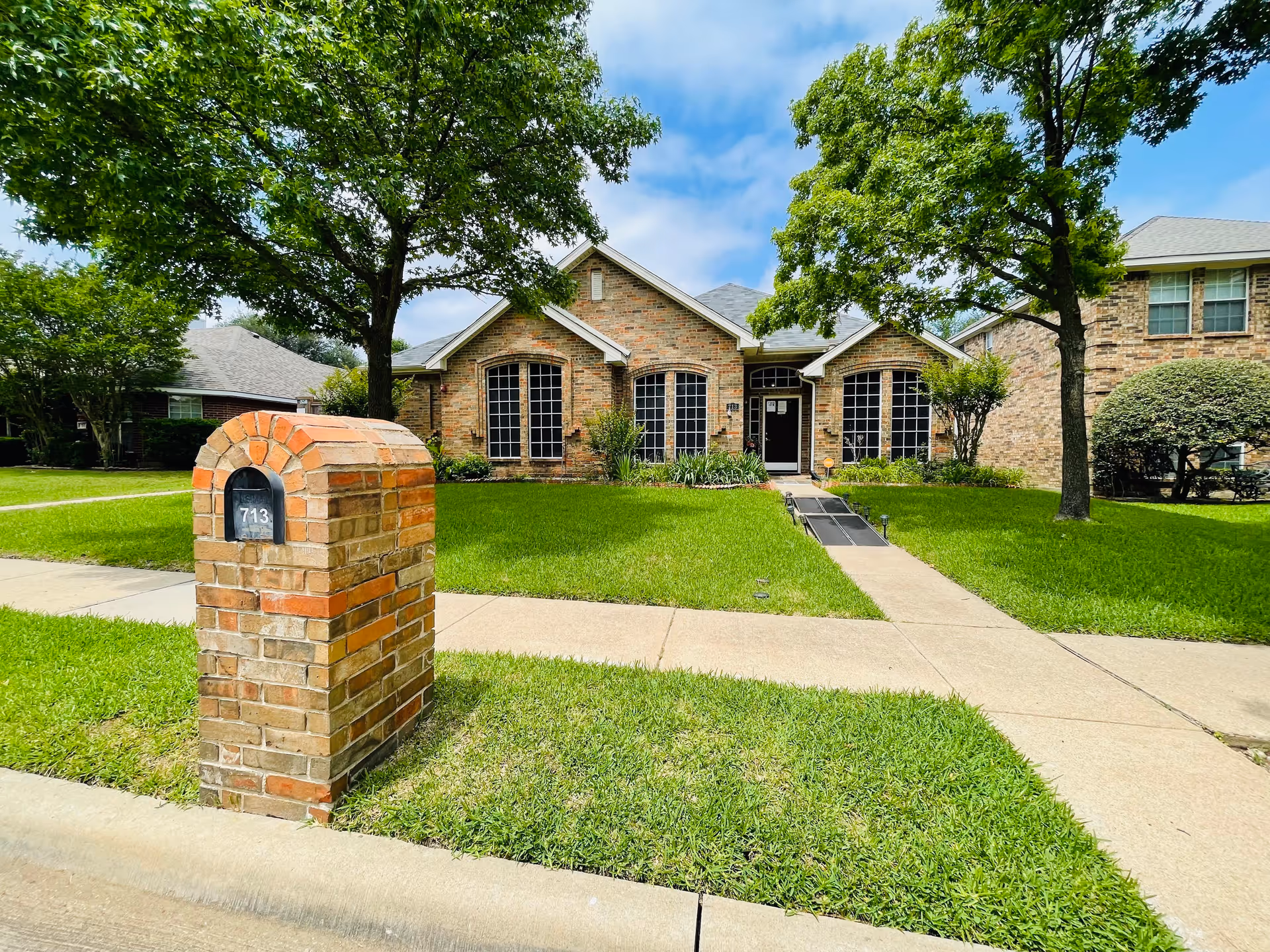 Front exterior view of a single-story brick house with large windows, a green lawn, a sidewalk, and a brick mailbox with the number 713. There are two trees on the lawn and a clear blue sky with some clouds.